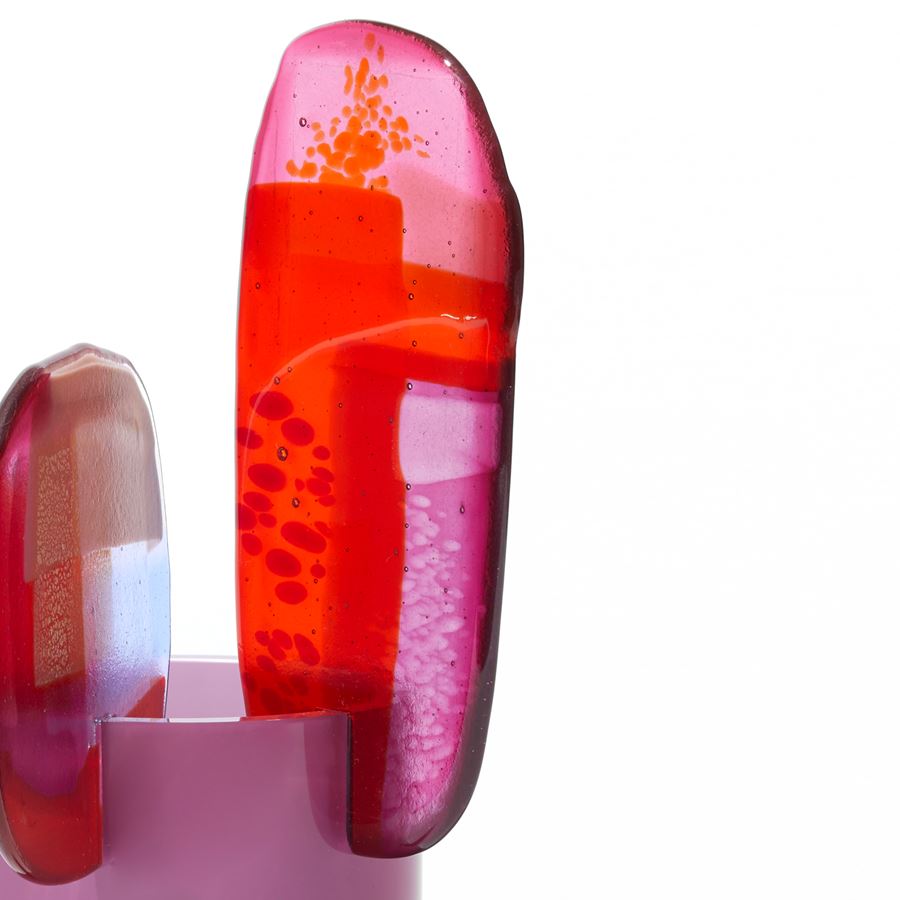 short opaque raspberry pink cylinder with three rounded different sized petals overlapping and perched on the top edge with abstract patterns in pink purple red and gold hand made from blown and fused glass